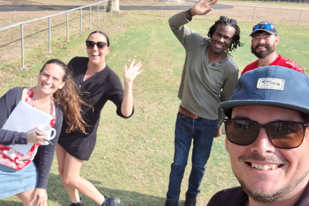 Group of five people standing outdoors all smiling at the camera. A man wearing sunglasses and a blue hat is taking the selfie photo, while the others wave at the camera.