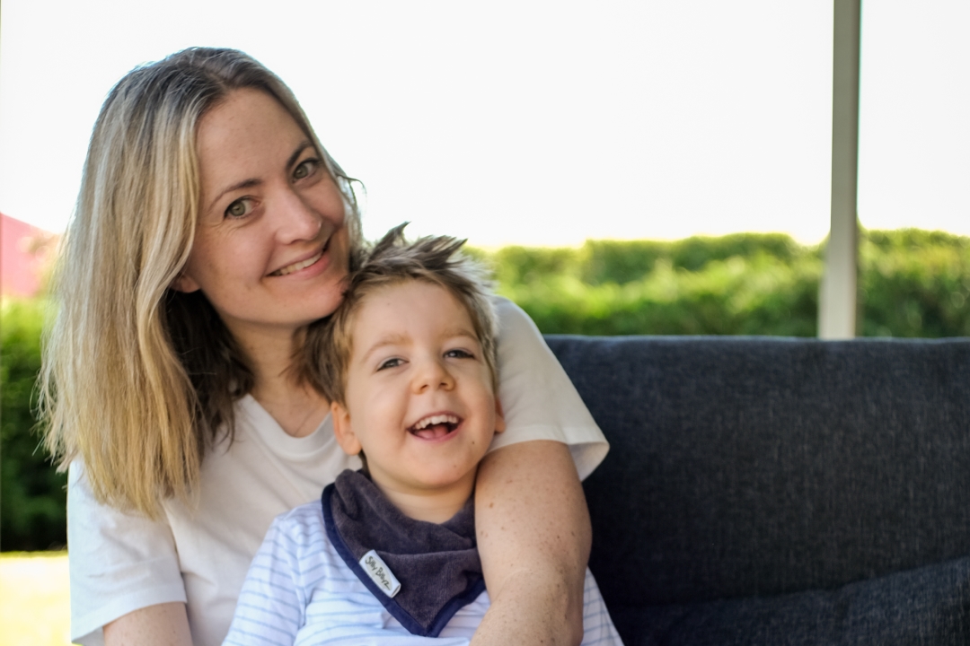 A little boy is sitting on his mums lap. She has with shoulder length blonde hair and wearing a white tshirt. They are sitting outside on a patio.