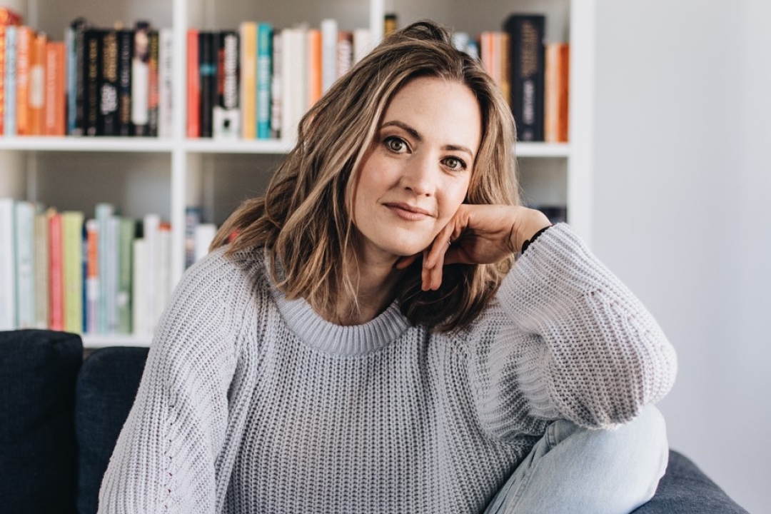 Melanie is wearing a grey jumper and is looking at the camera, resting her head on her hand.