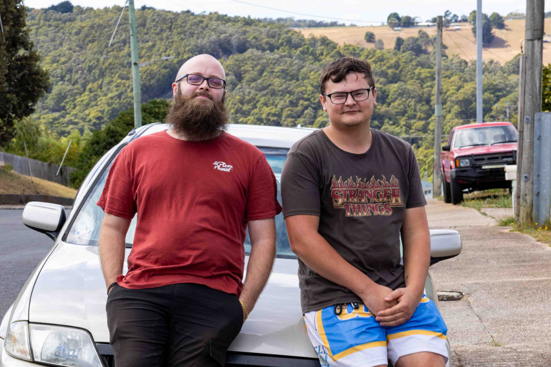 Jeremy is wearing a red t-shirt and black pants and Blade is wearing a black t-shirt with the words 'Stranger Things' on the front. Both are leaning on a car and smiling at the camera.