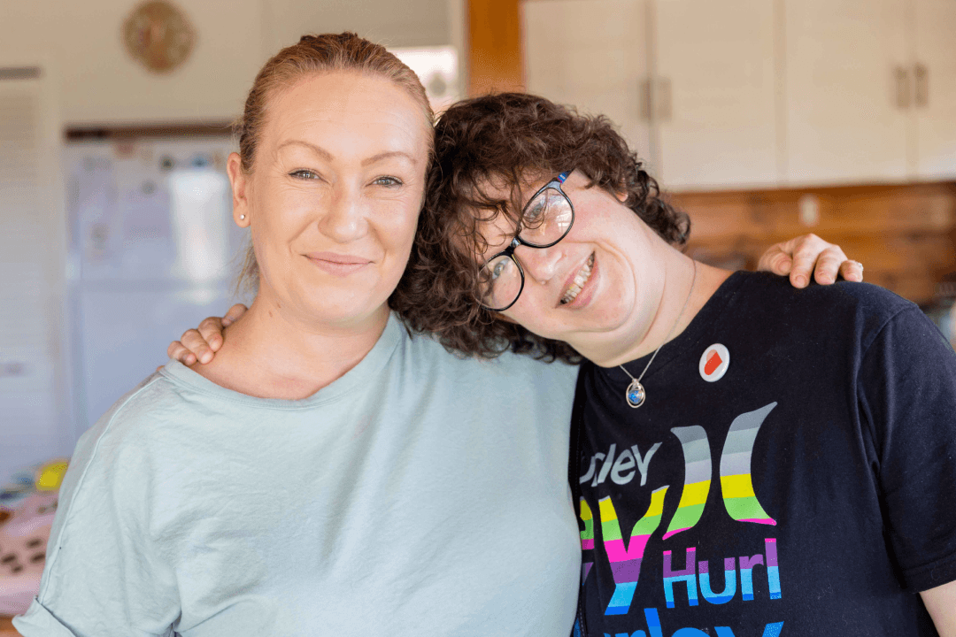 Isabella is wearing glasses and has short brown wavy hair. She has one arm around Janet's shoulder and is leaning her head on her shoulder. Janet has blonde hair tied back and is wearing a light green tshirt. Both are smiling at the camera.