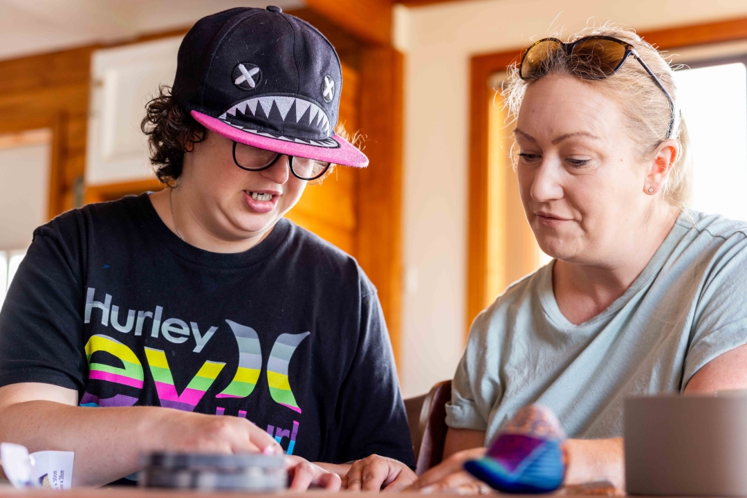 Isabella is wearing a black cap and tshirt. She is crafting and Janet is sitting beside her looking at what Isabella is doing.