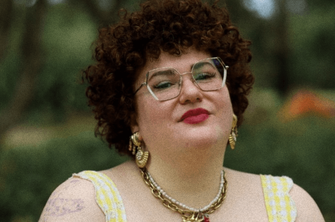 Woman with short dark curly hair, wearing glasses and a bright red lipstick She has her lips pursed and is looking at the camera