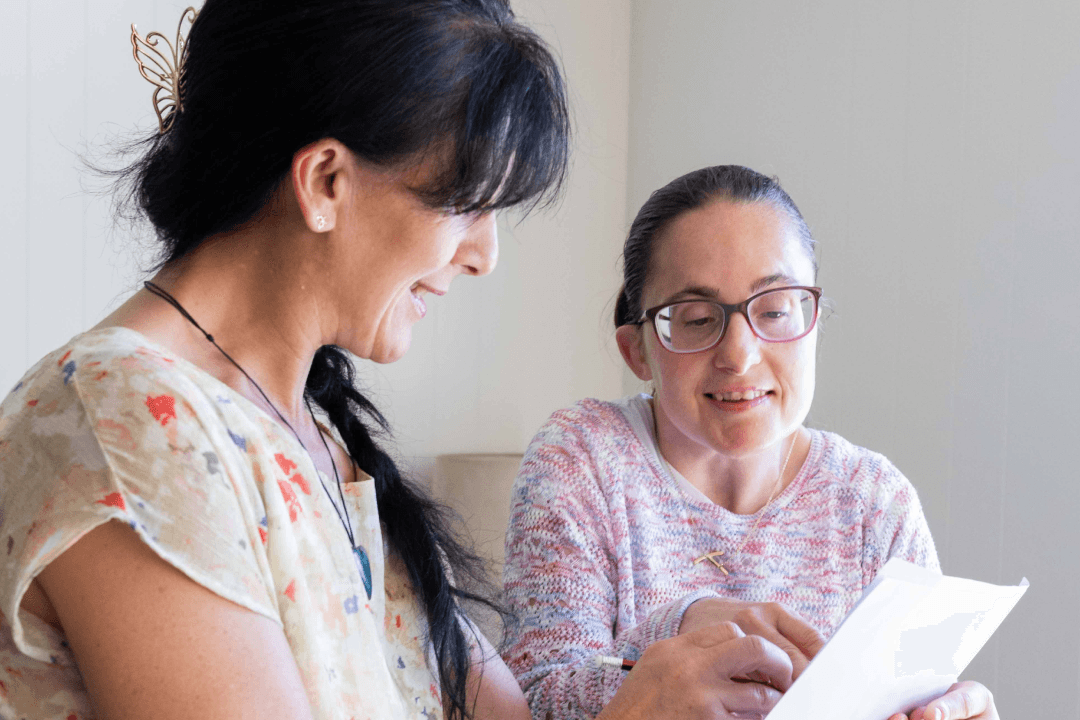 Angela is writing something on a piece of paper and Tiffany is sitting beside her. She is wearing glasses and knit jumper and she is looking at what Angela has written.