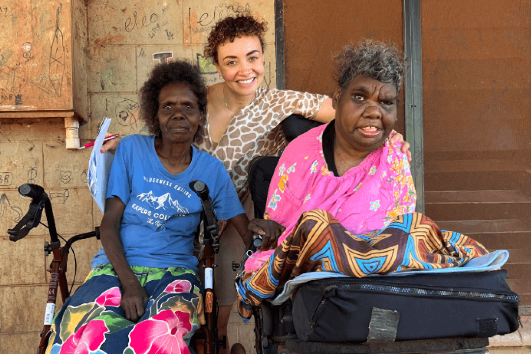 Veronica is wearing a pink top and is seated on the right in a wheelchair. Janita is seated to her left and is wearing a blue tshirt. Jane is standing up between them both.