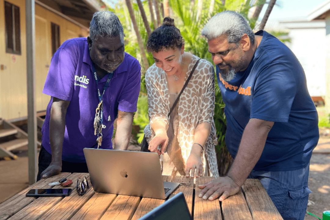 Jane is standing in between two First nations men. Everyon is looking and smiling at the laptop which is sitting on a wooden outdoor table.