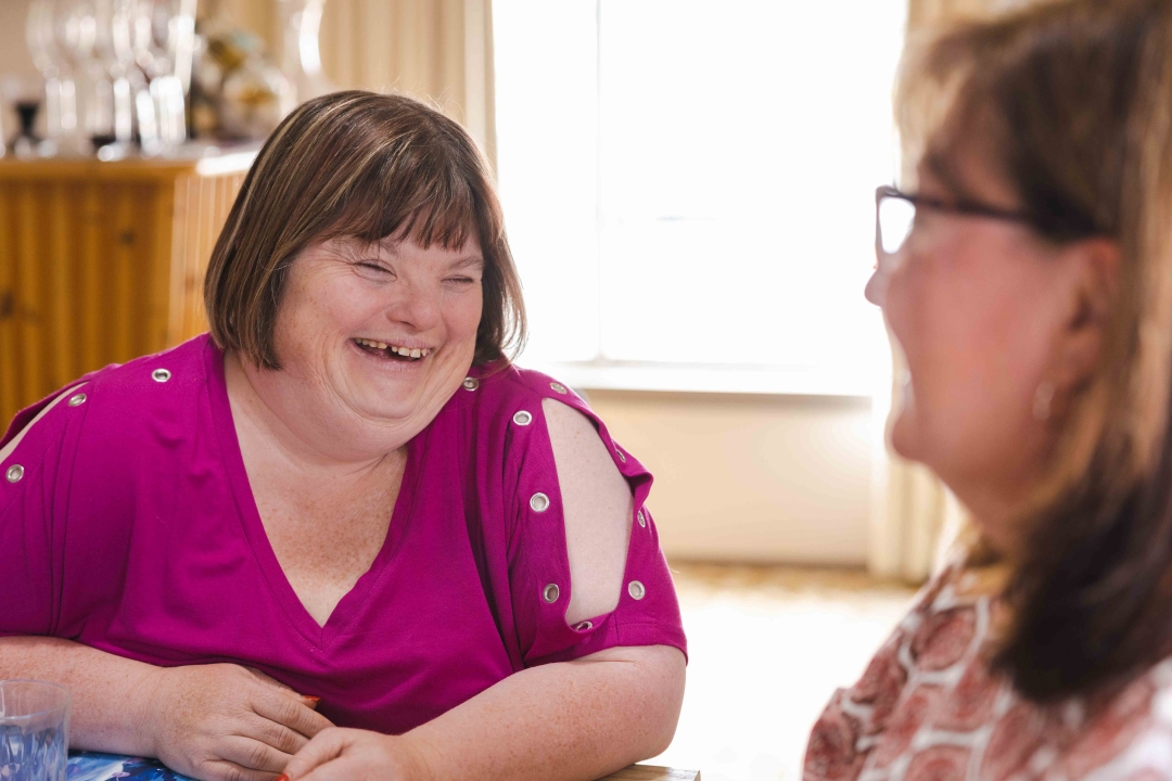 Woman with Down syndrome smiling at a woman with dark hair and glasses